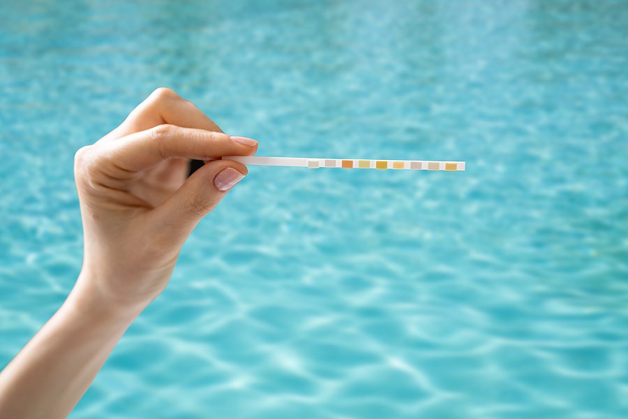 Hand holding a pool water test strip above clear blue water