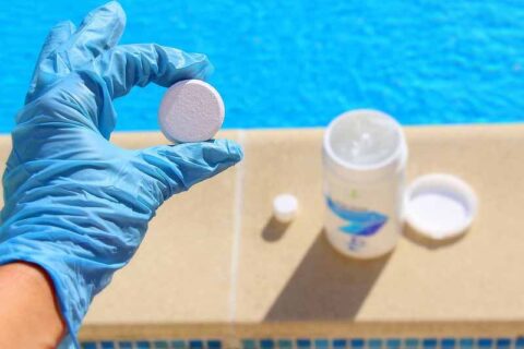 Gloved hand holding a pool chlorine tablet beside a swimming pool